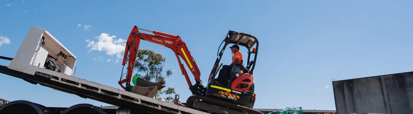 Man driving construction equipment loaded on a trailer truck at a worksite with safety barriers in the background.