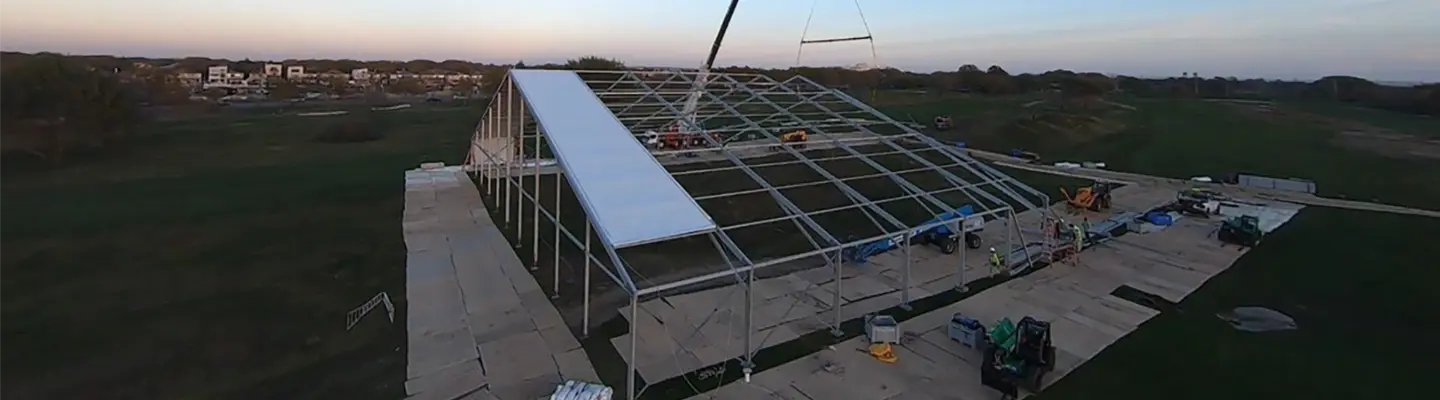 A crane installing a roof panel on a large temporary fabric structure at dusk in an open field