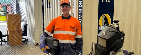 Person in hi-vis holding a welding helmet standing in front of a container.