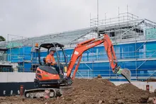 Man operating an excavator to excavate land near safety barriers on an active construction site.