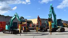 Row of excavators lined up at a construction site, with temporary safety barriers visible in the foreground.