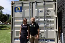 Two smiling company employees standing side by side in front of a storage container prepared for donation to a local community.